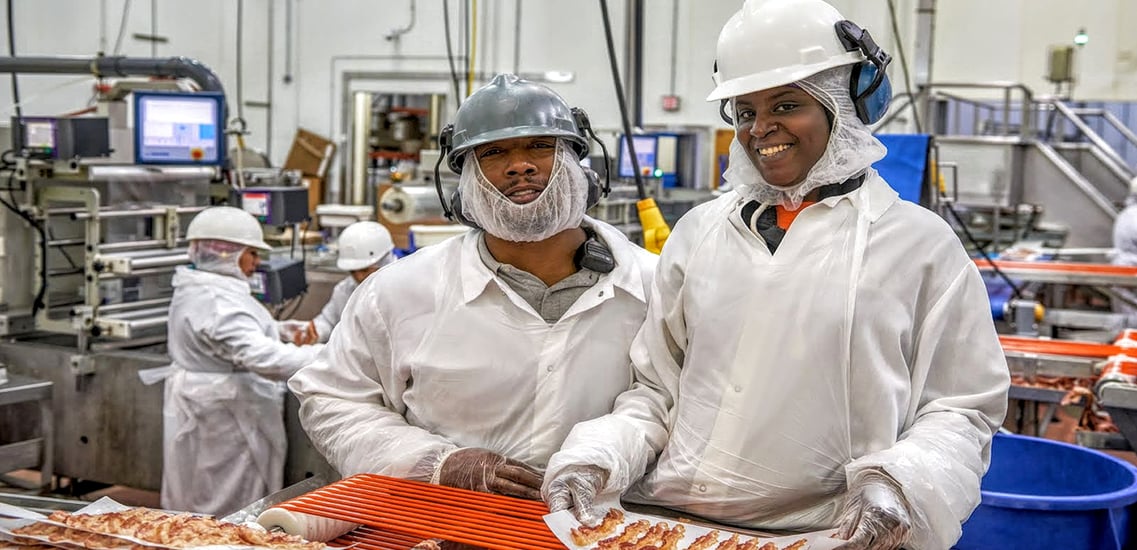 Two plant workers on the bacon line wearing white protection clothes smiling